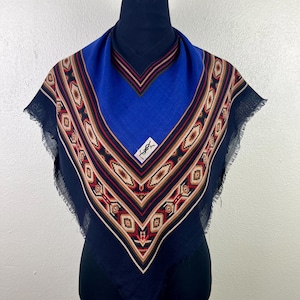 May include: A blue square scarf with a geometric pattern in red, brown, and beige. The scarf has a fringed edge and a small white label with a logo. The scarf is displayed on a black mannequin.