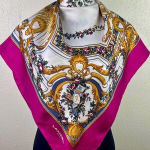 Puede incluir: Un pañuelo cuadrado de seda con un borde fucsia vibrante y un diseño floral y ornamental detallado. El pañuelo presenta detalles dorados, blancos y azules, con la marca "Lancel Paris" visible. También es visible un tocado a juego.