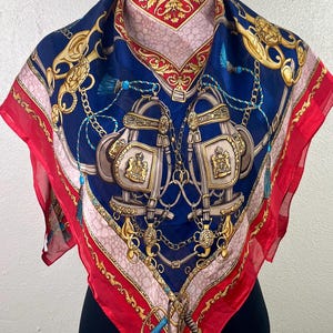 Puede incluir: Un pañuelo cuadrado de seda con fondo azul marino, con diseños dorados de temática ecuestre, cadenas y borlas. El pañuelo tiene un borde rojo y una pieza para el cuello con estampado rojo y dorado. El pañuelo se exhibe en un maniquí.