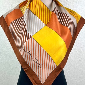 Puede incluir: Un pañuelo de seda con rayas y patrones geométricos marrones, naranjas, amarillos y blancos con el texto "Jean Cocteau" en escritura negra.