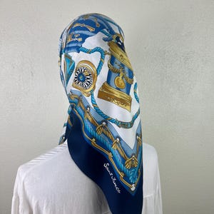 Puede incluir: Un pañuelo cuadrado de seda con temática náutica. Presenta un borde azul marino y un centro blanco con ilustraciones azules y doradas de brújulas, cuerdas y columnas decorativas. El texto "Saint James" está impreso en la esquina.