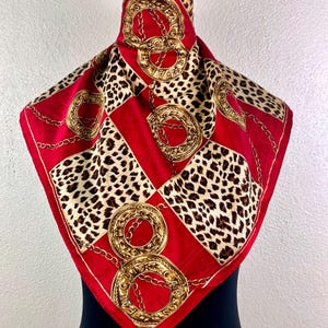 Puede incluir: Un pañuelo cuadrado con un diseño de cadena dorada, leopardo y rojo. El pañuelo está doblado, mostrando un patrón de cuadrados rojos, manchas de leopardo y eslabones de cadena dorados. El pañuelo es de material sedoso.