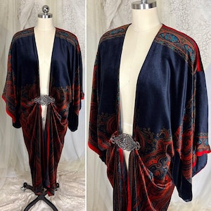 May include: A navy blue velvet kimono-style robe with a paisley pattern in red, orange, and teal. The robe features a decorative silver-tone clasp at the waist and red trim along the edges. The garment is displayed on a mannequin.