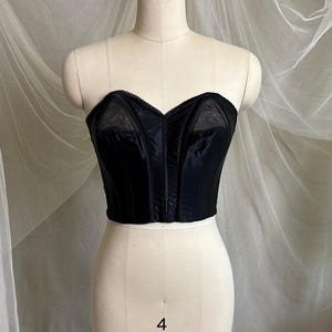May include: A black strapless corset top featuring a sweetheart neckline with sheer mesh panels at the upper bust. The garment is constructed with vertical boning channels and a satin-like finish, displayed on a mannequin with the number 4 visible.