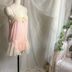 May include: A pale pink camisole bodysuit featuring thin spaghetti straps, a cream-colored lace bodice overlay with a small central bow, and a ruffled cream-colored lace trim along the leg openings, displayed on a dress form mannequin.