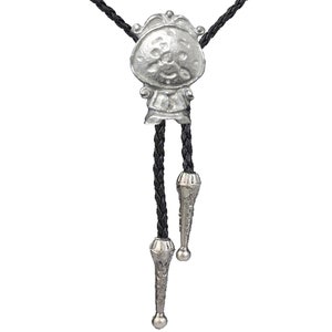 Może przedstawiać: Srebrny krawat bolo z zawieszką w postaci postaci z kreskówek. Postać ma okrągłą głowę z uśmiechającą się twarzą i koroną. Krawat jest wykonany ze splecionego czarnego skóry.