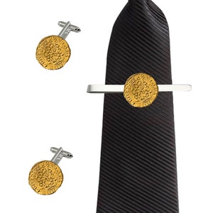 Puede incluir: Un juego de gemelos y un pasador de corbata dorados con un diseño circular. El pasador de corbata está sujeto a una corbata negra a rayas.
