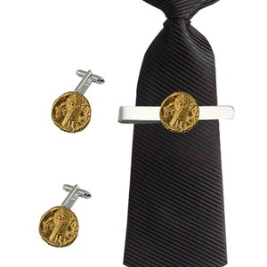 Puede incluir: Un juego de gemelos dorados y un pasador de corbata con una réplica de una antigua moneda romana. El pasador de corbata es plateado y la corbata es negra.