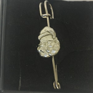 May include: Silver-toned brooch featuring a detailed Santa Claus head design. The brooch is attached to a safety pin.