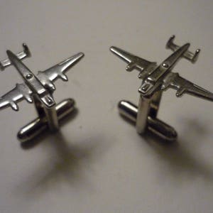May include: A pair of silver cufflinks featuring a detailed airplane design. The cufflinks are made of metal and have a polished finish.