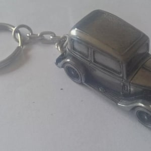 May include: A silver metal keychain featuring a vintage car model. The car is a sedan with a rounded body and a detailed front grille.