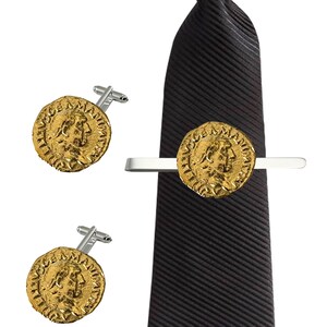 Puede incluir: Un juego de gemelos y pasador de corbata de oro con una réplica de una antigua moneda romana. El pasador de corbata y los gemelos son plateados. El pasador de corbata está sujeto a una corbata negra.