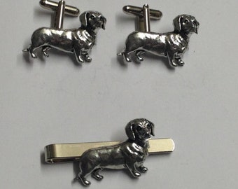 Puede incluir: Un juego de gemelos de plata con forma de dachshund y un clip para corbata. El dachshund es una raza de perro pequeña con patas cortas y cuerpo largo.