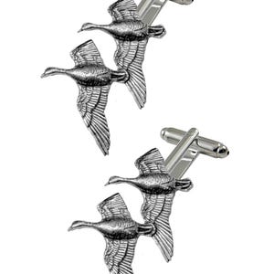 May include: A pair of silver cufflinks featuring a design of two geese in flight. The geese are depicted in a realistic style with detailed wings and feathers.