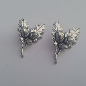 Könnte beinhalten: Ein Paar silberfarbene Ohrringe mit Eichel und Eichenblatt. Die Ohrringe sind aus Metall und haben ein detailliertes Design.