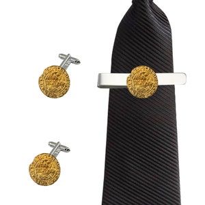 Puede incluir: Un juego de gemelos y un pasador de corbata dorados con un diseño circular. Los gemelos y el pasador de corbata son plateados y tienen un acabado pulido. El pasador de corbata está sujeto a una corbata negra.