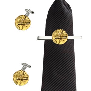 Puede incluir: Un juego de gemelos dorados y un clip para corbata con una réplica de una moneda antigua. La moneda tiene escritura hebrea y una representación de un edificio. El clip para corbata está unido a una corbata negra.