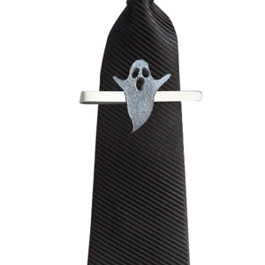 Puede incluir: Una corbata negra con un clip de corbata de fantasma. El fantasma es plateado y tiene una cara blanca con la boca abierta en un grito.