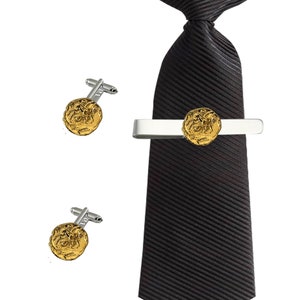 Puede incluir: Un juego de gemelos dorados y un pasador de corbata con un diseño detallado de una moneda romana. El pasador de corbata está unido a una corbata negra a rayas.