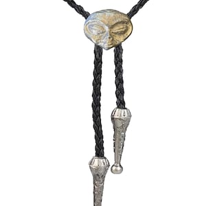Peut inclure: Un bolo tie en cuir tressé noir avec un pendentif tête d'extraterrestre argenté et doré et des embouts argentés.