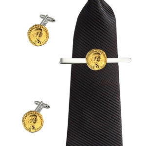 Puede incluir: Una corbata negra con un clip de corbata de moneda de oro y gemelos a juego de moneda de oro. La moneda presenta un perfil de una persona con texto alrededor del borde.