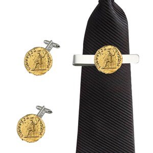 Puede incluir: Una corbata negra con un clip de corbata plateado que presenta una moneda de oro con letras romanas. El conjunto también incluye un par de gemelos con el mismo diseño de moneda de oro.