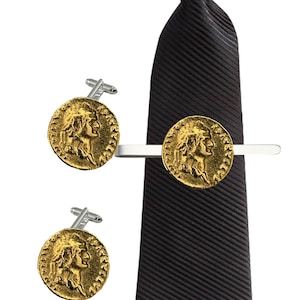 Puede incluir: Un juego de gemelos y un pasador de corbata dorados con un diseño de moneda romana. El pasador de corbata está unido a una corbata negra a rayas.