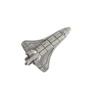 Op de afbeelding: Een zilverkleurige ruimteshuttle-speld. De speld heeft gedetailleerde panelen en een getextureerd oppervlak, met een kleine motor aan de achterkant. De shuttle staat iets schuin, wat het aerodynamische ontwerp laat zien.