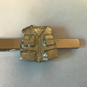 May include: A silver tie clip featuring a detailed design of a vest with pockets.