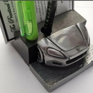 May include: A silver car desk organiser with a pen and a green highlighter. The organiser has a wooden base with a brushed metal finish. The text "The Practical Glasses & Pen Holder" is visible on a card.