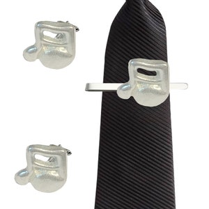May include: A set of silver musical note cufflinks and a tie clip. The tie clip is attached to a dark brown striped tie.