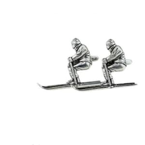 May include: A pair of silver cufflinks featuring a skier on skis. The skier is wearing a hat and jacket.