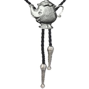 Może przedstawiać: Srebrny krawat bolo w kształcie czajnika z czarnymi plecionymi sznurkami ze skóry. Czajnik ma szczegółowy wzór i srebrne wykończenie.