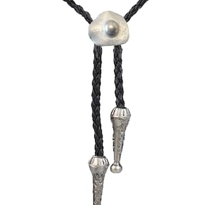 May include: A black braided bolo tie with silver-toned tips and a silver-toned heart-shaped pendant with a round bead in the centre.
