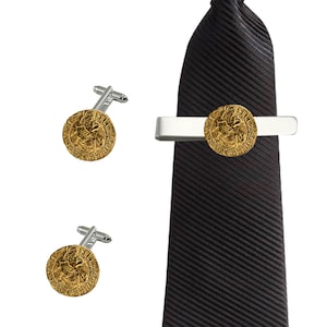 Puede incluir: Un juego de gemelos dorados y un pasador de corbata con un diseño redondo con una imagen detallada. El pasador de corbata está unido a una corbata negra.