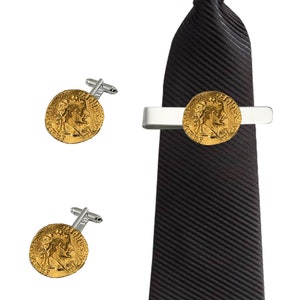 Puede incluir: Un juego de gemelos y un pasador de corbata dorados con un diseño de moneda romana. El pasador de corbata está sujeto a una corbata negra.