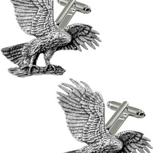 Könnte beinhalten: Ein Paar silberne Manschettenknöpfe mit einem detaillierten, geformten Design eines Adlers mit ausgebreiteten Flügeln. Der Adler sitzt auf einem Ast und hat ein realistisches Aussehen.