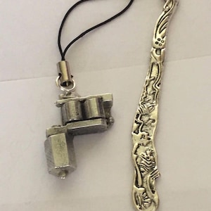 Puede incluir: Un marcapáginas de plata con un diseño de dragón y un amuleto de plata en forma de máquina de tatuaje. El marcapáginas tiene una cuerda negra.