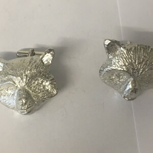 May include: A pair of silver cufflinks featuring a raccoon head design. The cufflinks are detailed with a textured fur pattern.
