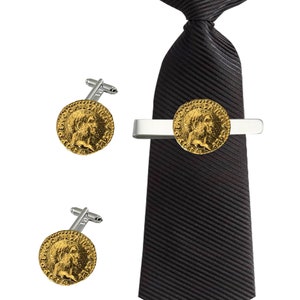 Puede incluir: Un juego de gemelos y un clip para corbata de color dorado con un diseño de moneda romana. El clip para corbata está sujeto a una corbata negra.