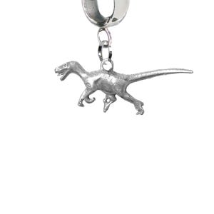 May include: A silver charm depicting a raptor dinosaur. The charm has a loop for attaching to a bracelet or necklace. The dinosaur is shown in a running pose, with detailed features. The charm is made of a silver-coloured metal.