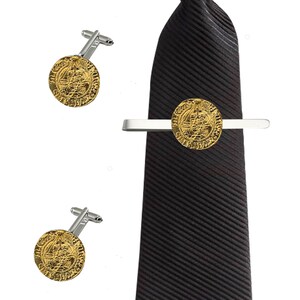 Puede incluir: Un juego de gemelos y pasador de corbata de oro con un diseño de moneda redonda. El pasador de corbata es de plata con un diseño de moneda de oro. Los gemelos son de plata con diseños de monedas de oro. El pasador de corbata y los gemelos están en una corbata de rayas negras.