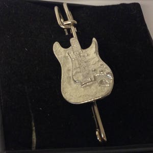 May include: Silver-toned guitar-shaped tie clip. The clip features a detailed depiction of an electric guitar, including the body, neck, and strings. The tie clip is set against a black velvet background.