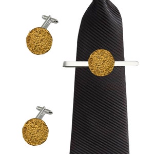 Puede incluir: Un juego de gemelos y un pasador de corbata dorados con un diseño circular con detalles intrincados. El pasador de corbata está unido a una corbata negra a rayas.