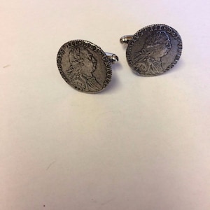 May include: A pair of silver cufflinks featuring a detailed design of a person's profile. The cufflinks are circular and have a raised edge.