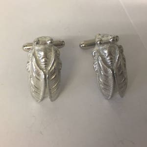 May include: A pair of silver-colored cufflinks, each shaped like a cicada insect. The cufflinks have detailed wings and body features, with a post for securing them to a shirt cuff. The background is a plain white surface.
