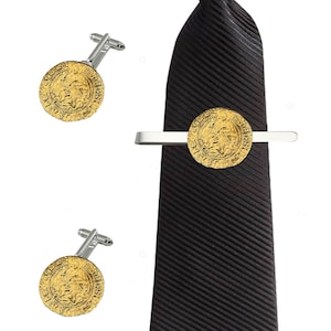 Puede incluir: Una corbata negra con un broche de corbata dorado y un par de gemelos dorados. El broche de corbata y los gemelos tienen un diseño redondo con una imagen en relieve de una persona.