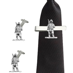 Puede incluir: Un juego de gemelos y pinza de corbata de plata con una figura en miniatura de una persona sosteniendo un rifle. La pinza de corbata está unida a una corbata negra.