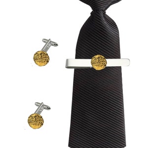 Puede incluir: Un juego de gemelos dorados y un pasador de corbata con un diseño decorativo. El pasador de corbata está sujeto a una corbata negra a rayas.