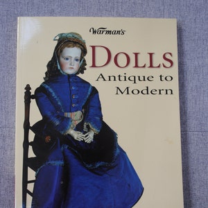 May include: A book titled "Warman's Dolls Antique to Modern" by Mark F. Moran. The cover features a vintage doll wearing a blue dress and sitting on a wooden chair. The book is an identification and price guide.